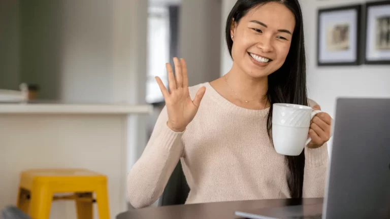 A woman sits at a table with a laptop, smiling and waving with one hand while holding a white mug in the other, appearing to be on a video call in a cozy, home setting.
