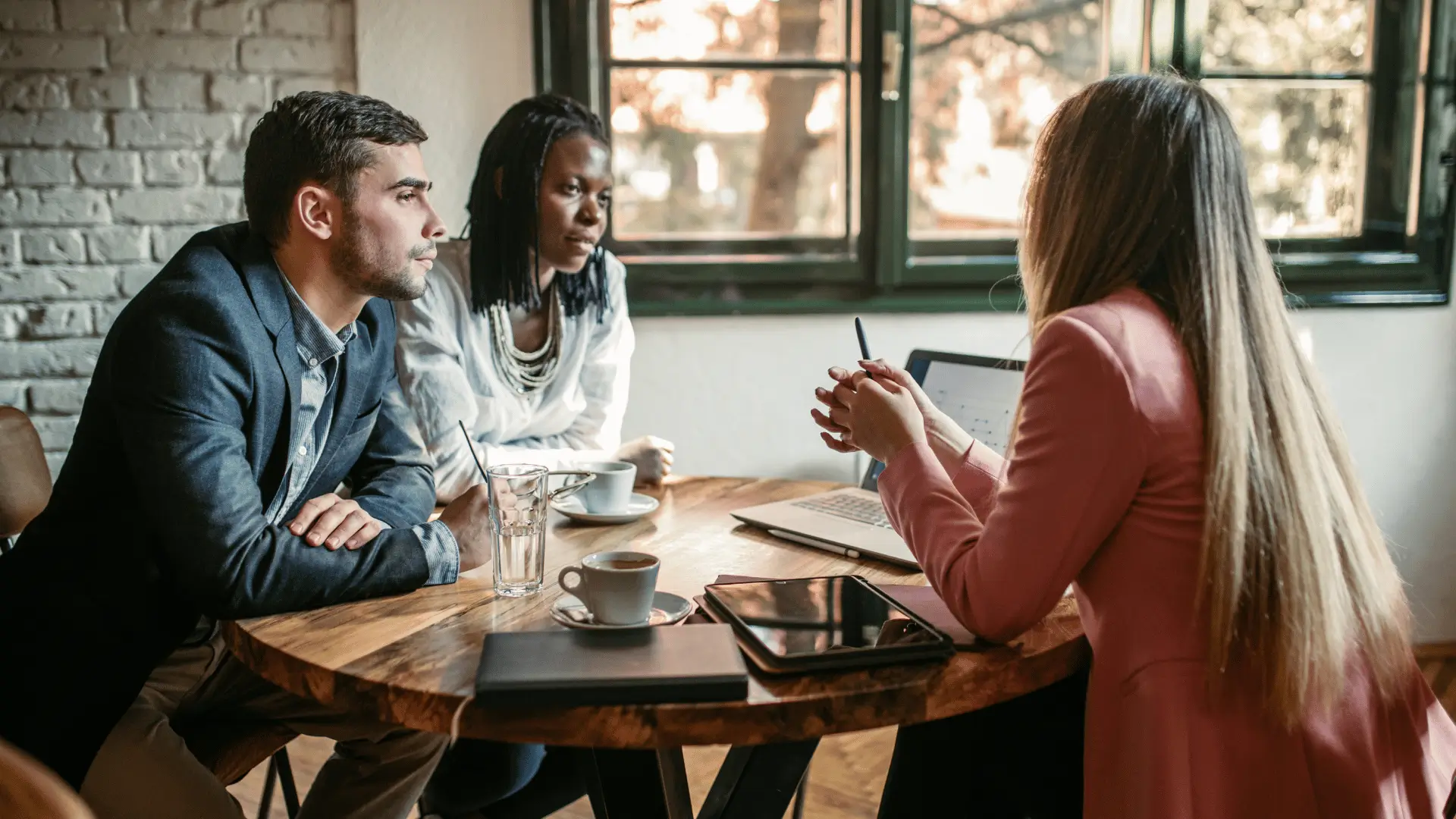Three people sit around a round wooden table having a discussion. Two listen attentively while one speaks. A laptop, tablet, notebook, and coffee cups are on the table. Sunlight comes through windows in the background.