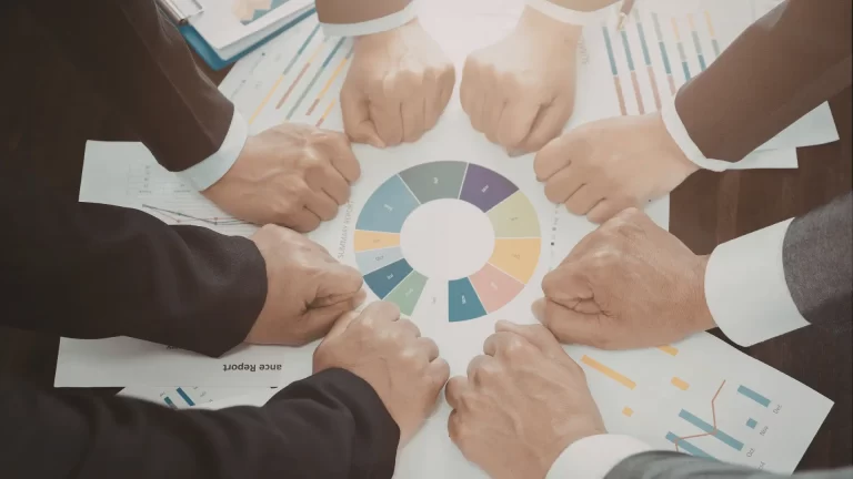 Eight people in business attire place their fists together over a circular business chart on a table covered with financial documents and reports, symbolizing teamwork and unity.