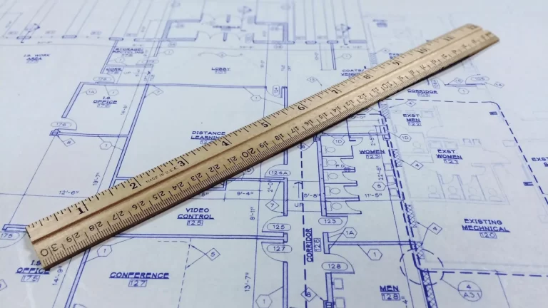 A wooden ruler is placed diagonally on a detailed architectural blueprint with various rooms, labels, and measurements visible in blue ink.