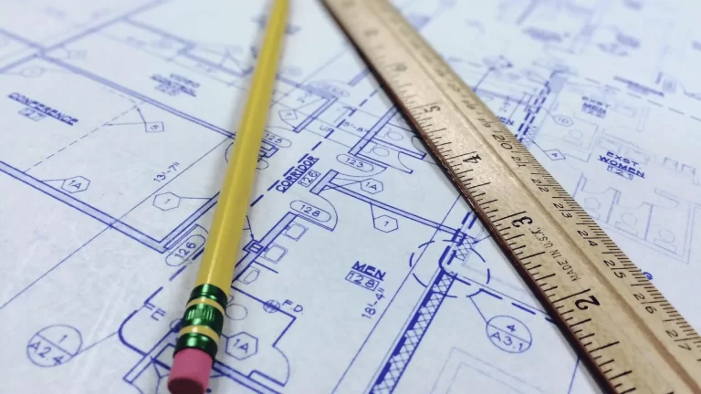 A yellow pencil and a metal ruler are placed on top of blue architectural floor plans, showing detailed room layouts and measurements.