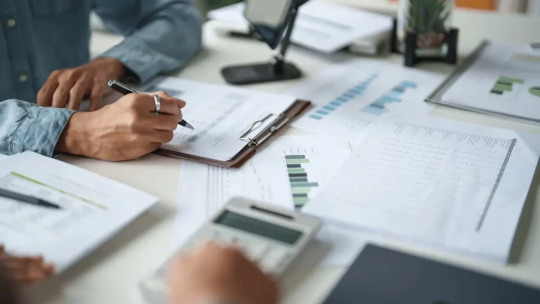 A person reviews and analyzes financial documents and graphs at a desk with scattered papers, a clipboard, a calculator, and a small potted plant.