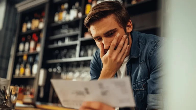 A man in a denim shirt looks concerned as he examines a bill or document in his hand, standing in front of shelves filled with bottles in a dimly lit bar or restaurant.