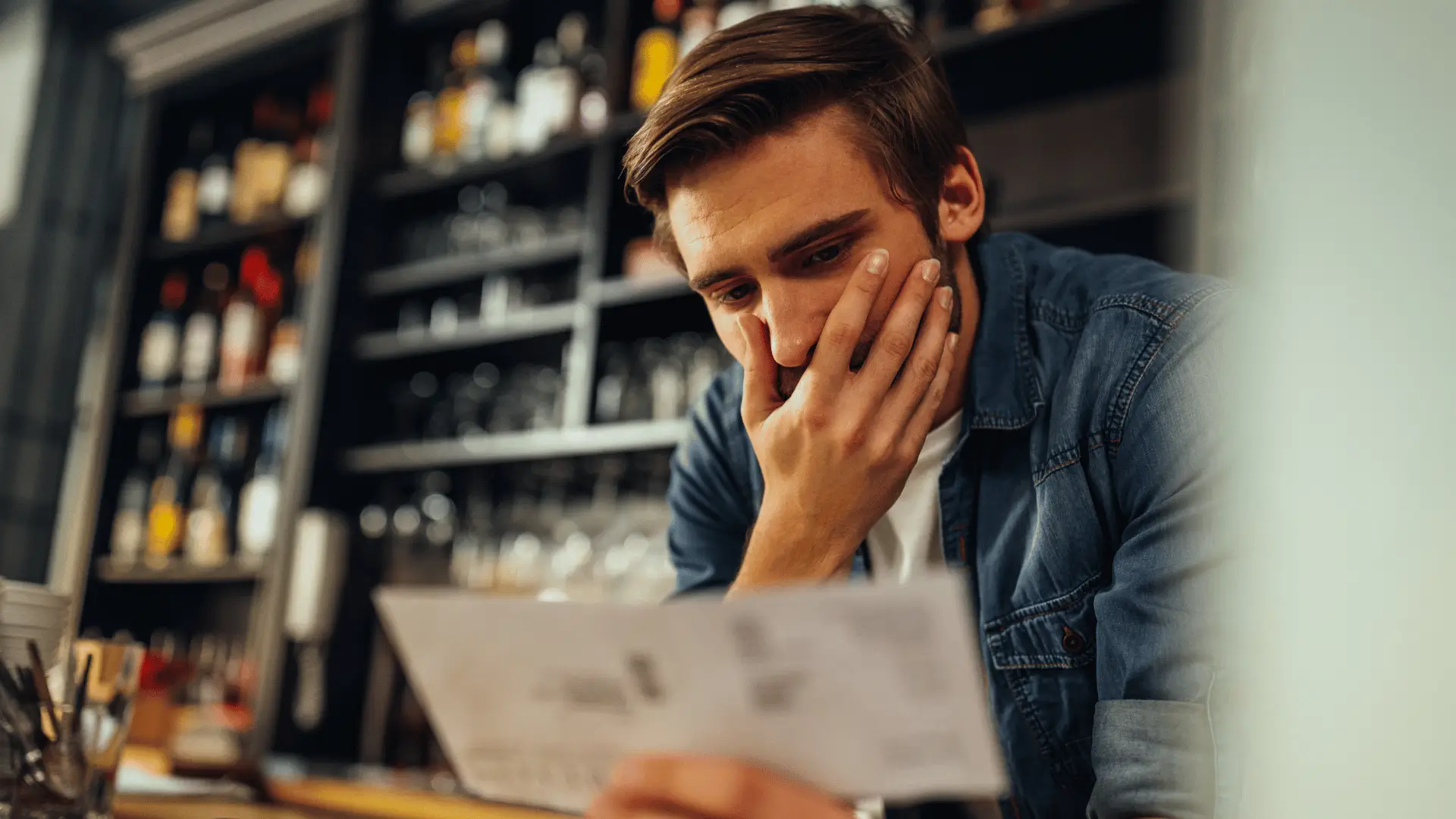 A man in a denim shirt looks concerned as he examines a bill or document in his hand, standing in front of shelves filled with bottles in a dimly lit bar or restaurant.