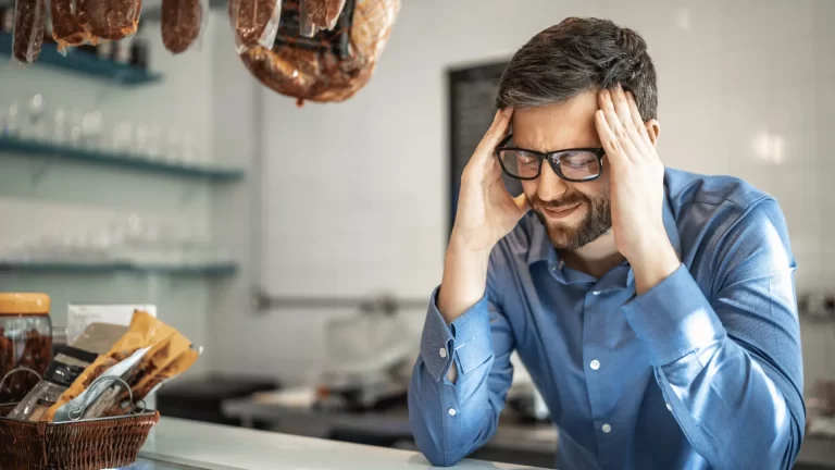A man in a blue shirt and glasses sits at a counter, holding his head with both hands and appears stressed or frustrated. Shelves, sausages, and baskets with packages are visible in the background.